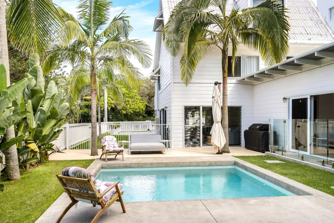 Piscina exterior junto a una casa blanca, rodeada de palmeras y asientos de patio.