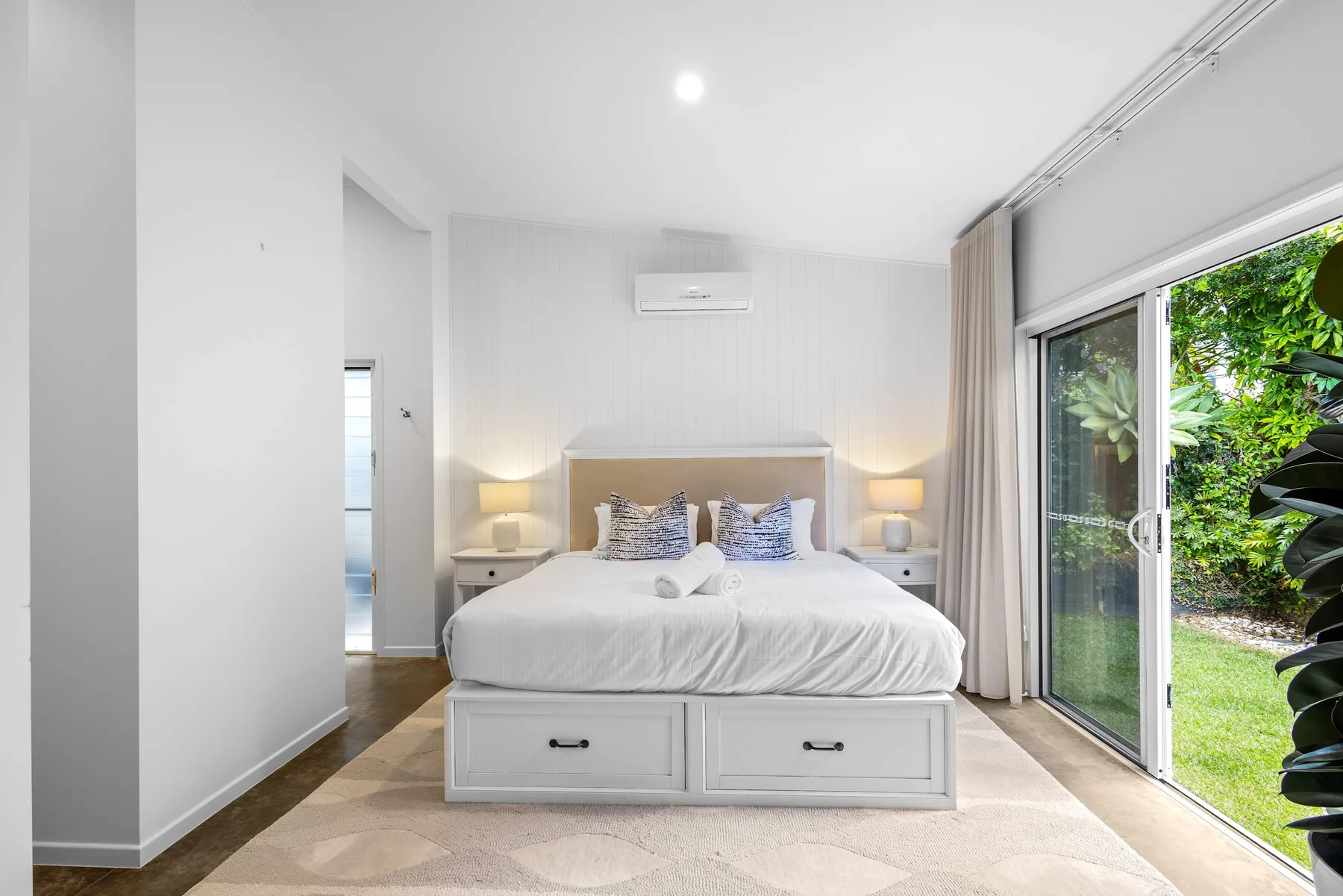 Un dormitorio luminoso tiene una cama entre mesitas de noche y una gran puerta de vidrio.