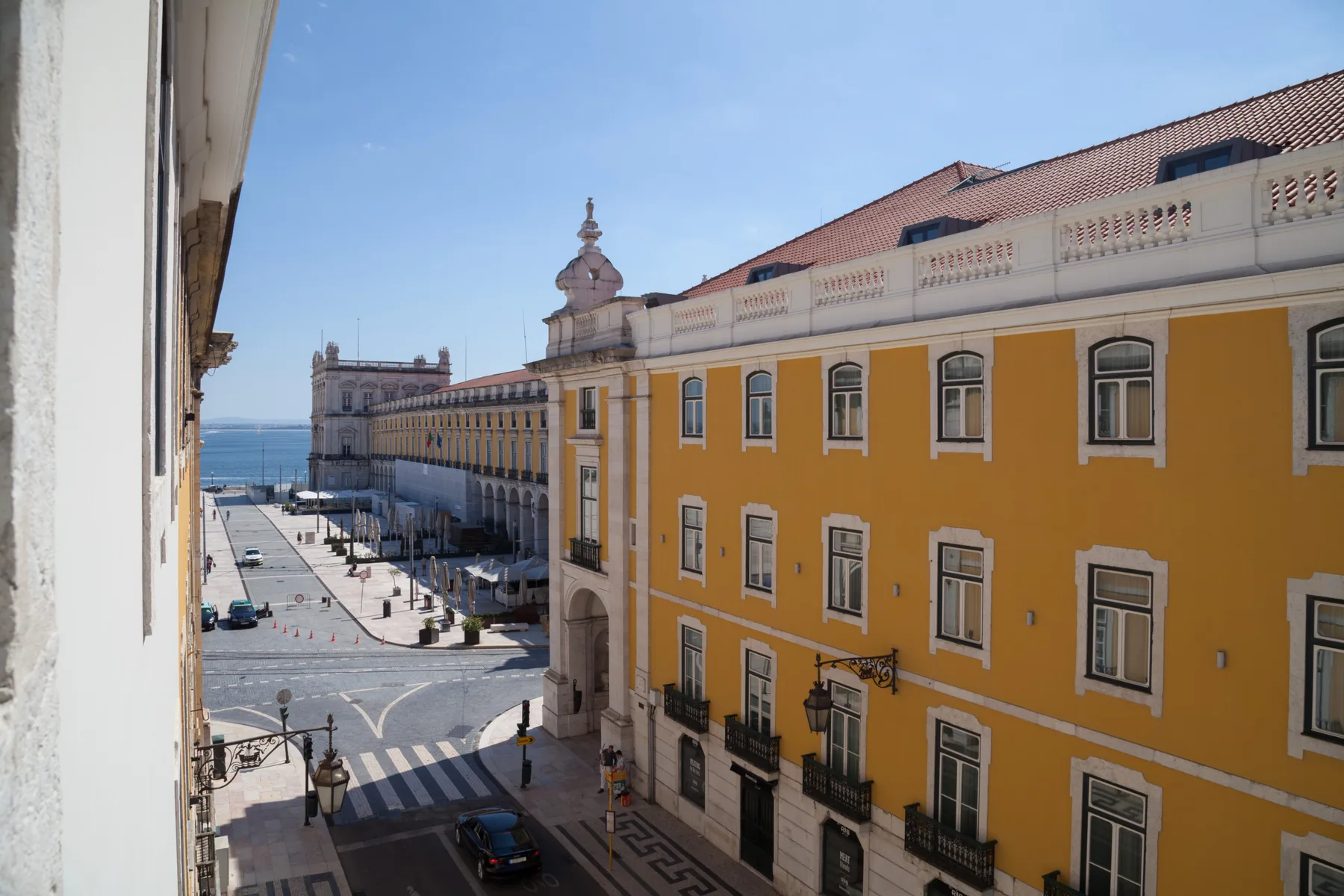 Una plaza urbana junto a un largo edificio amarillo, con el paseo marítimo al fondo.