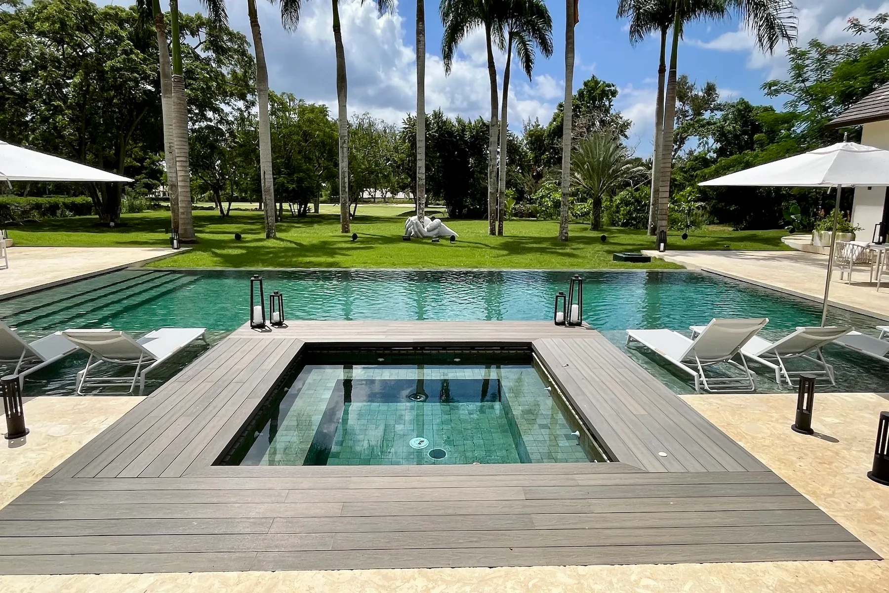 Una piscina al aire libre y un jacuzzi integrado están rodeados por una amplia terraza de madera y palmeras.