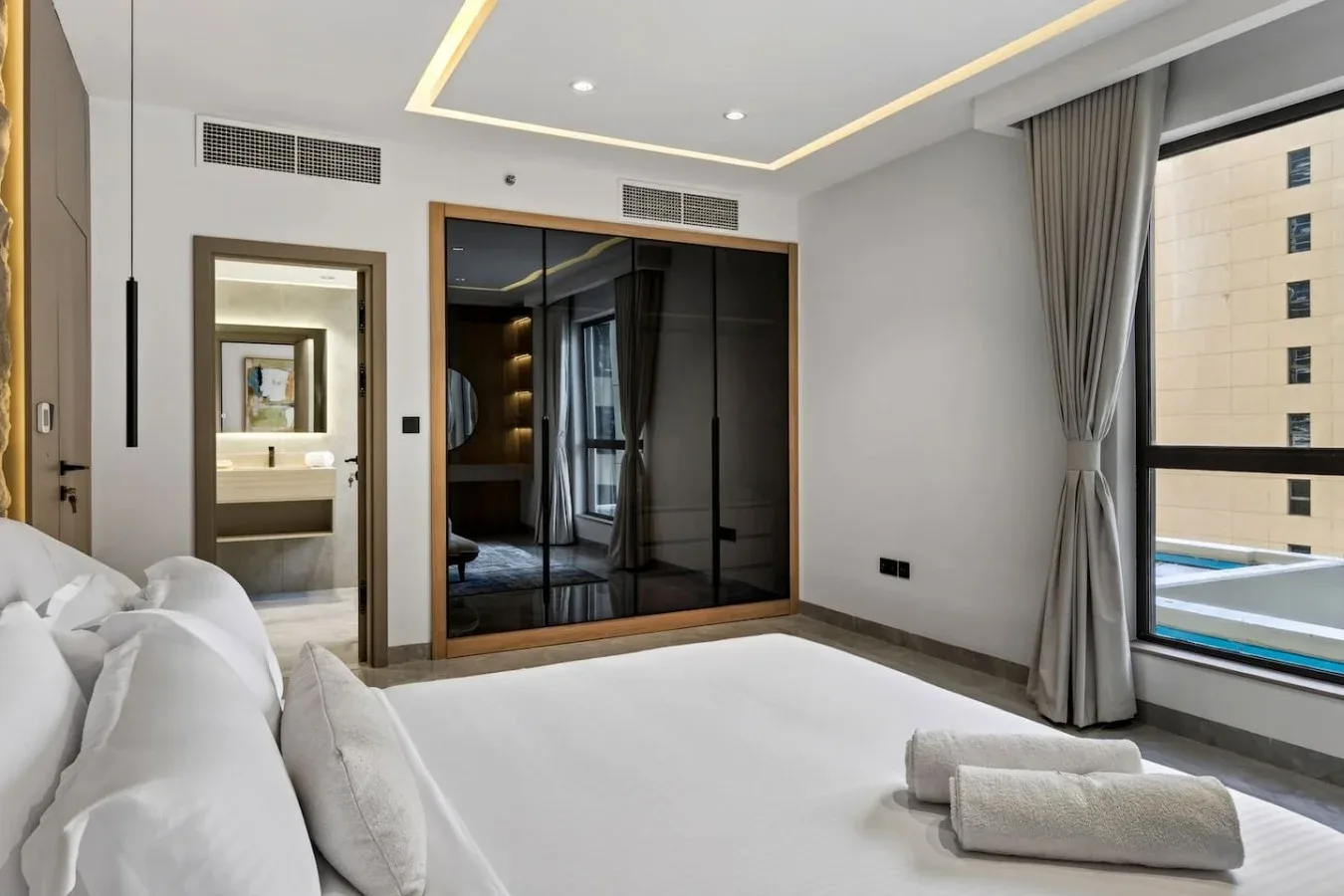 A bright bedroom with white bedding and dark mirrored wardrobe doors.