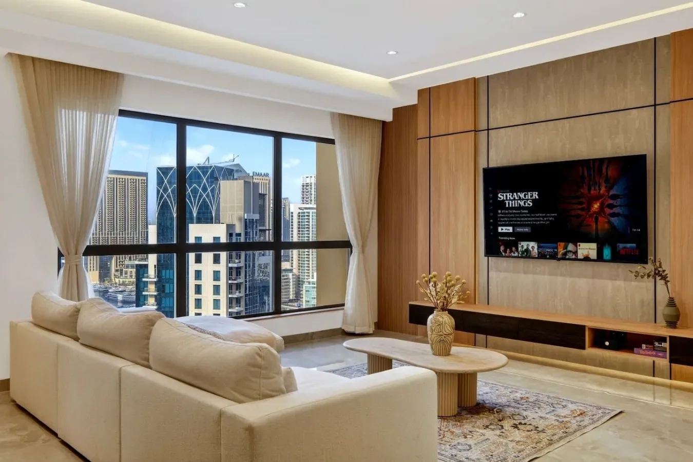 A living room with a cream seating area, a coffee table, and large windows overlooking city buildings.