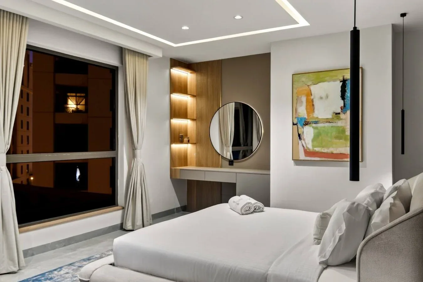 A modern bedroom with a neatly made bed and a round mirror above the vanity.