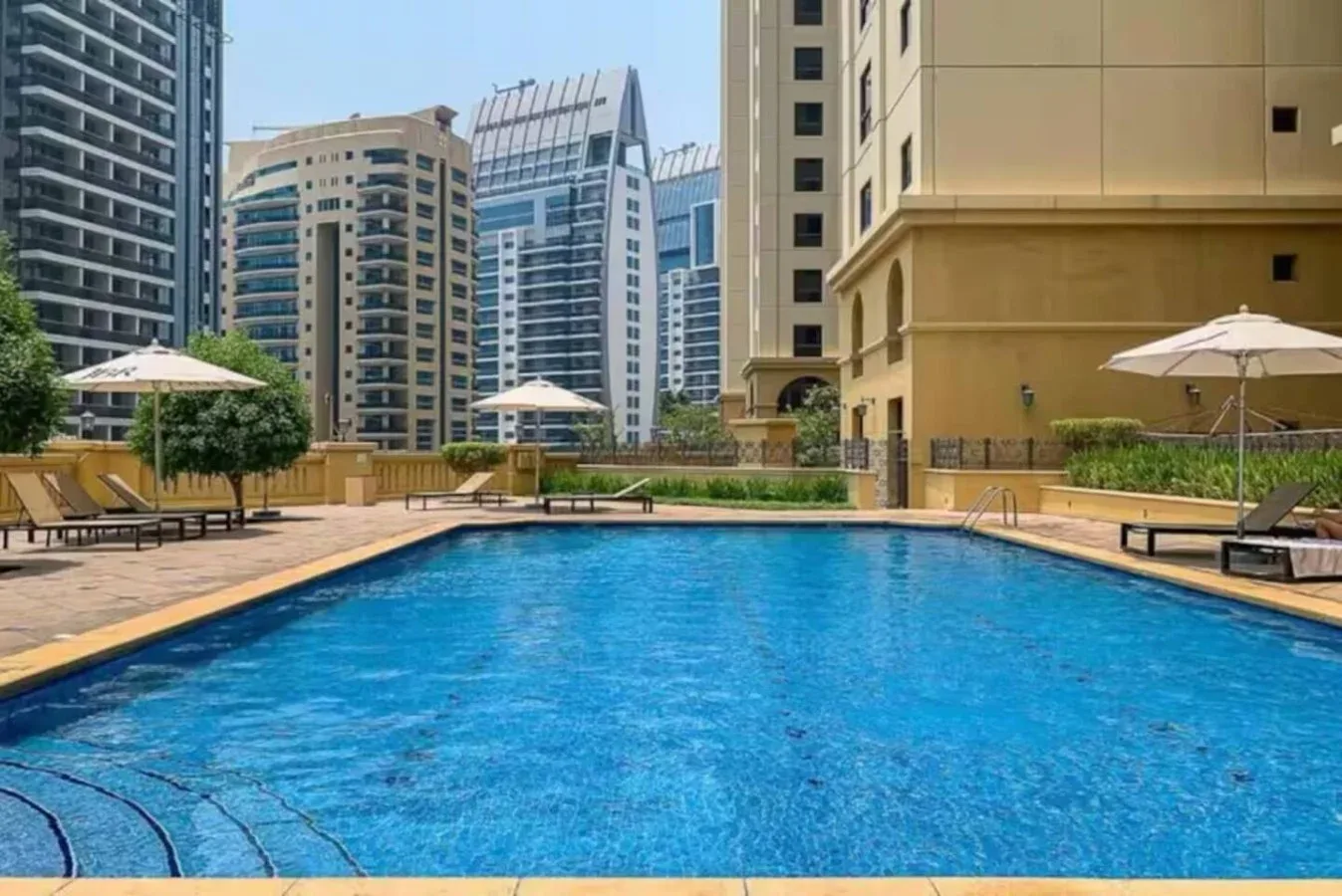 An outdoor swimming pool sits among tall buildings, with umbrellas and lounge seating along the deck.