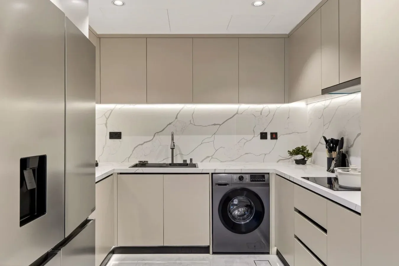 A modern kitchen with pale cabinets, a washing machine, and a stainless-steel refrigerator.
