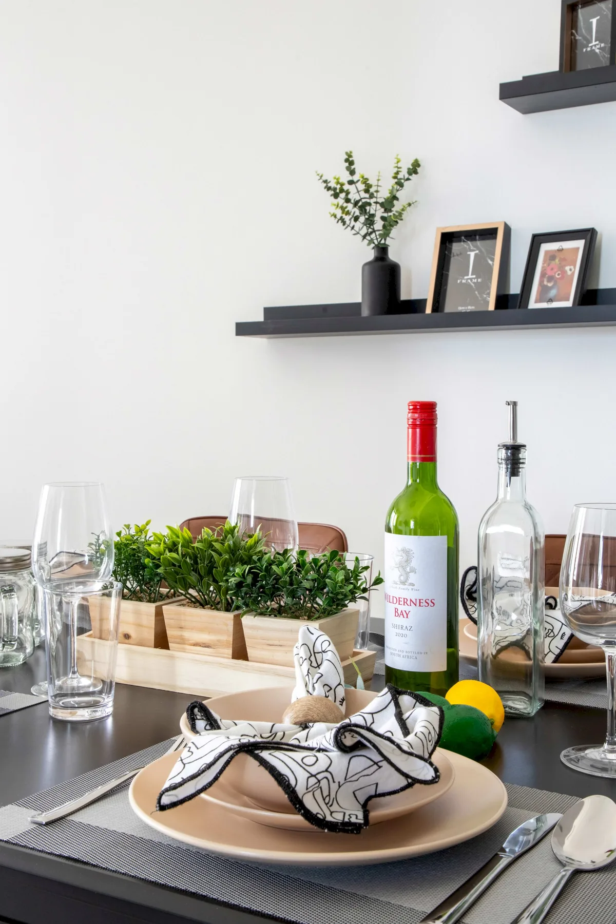 Una mesa de comedor preparada con platos, copas, botellas y una planta pequeña.