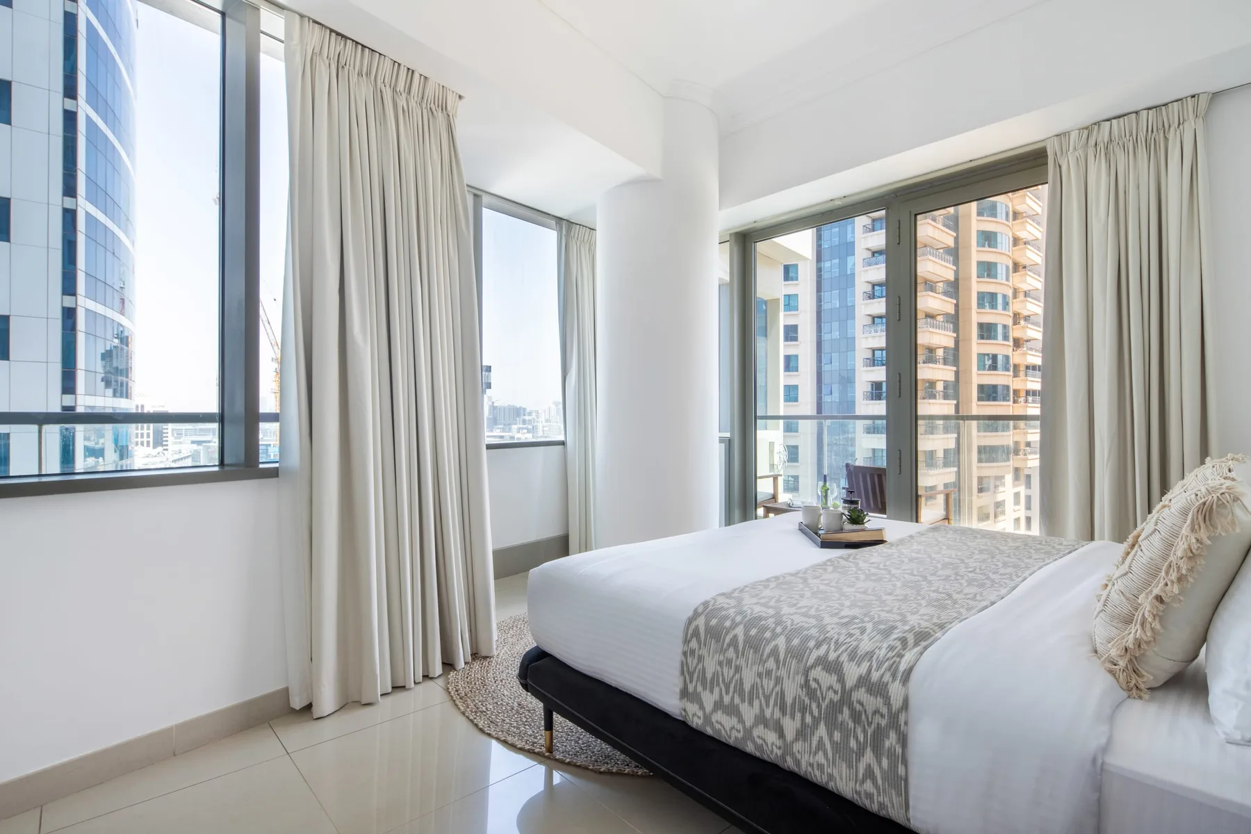 Un dormitorio luminoso con una cama junto a grandes ventanas que dan a edificios cercanos.