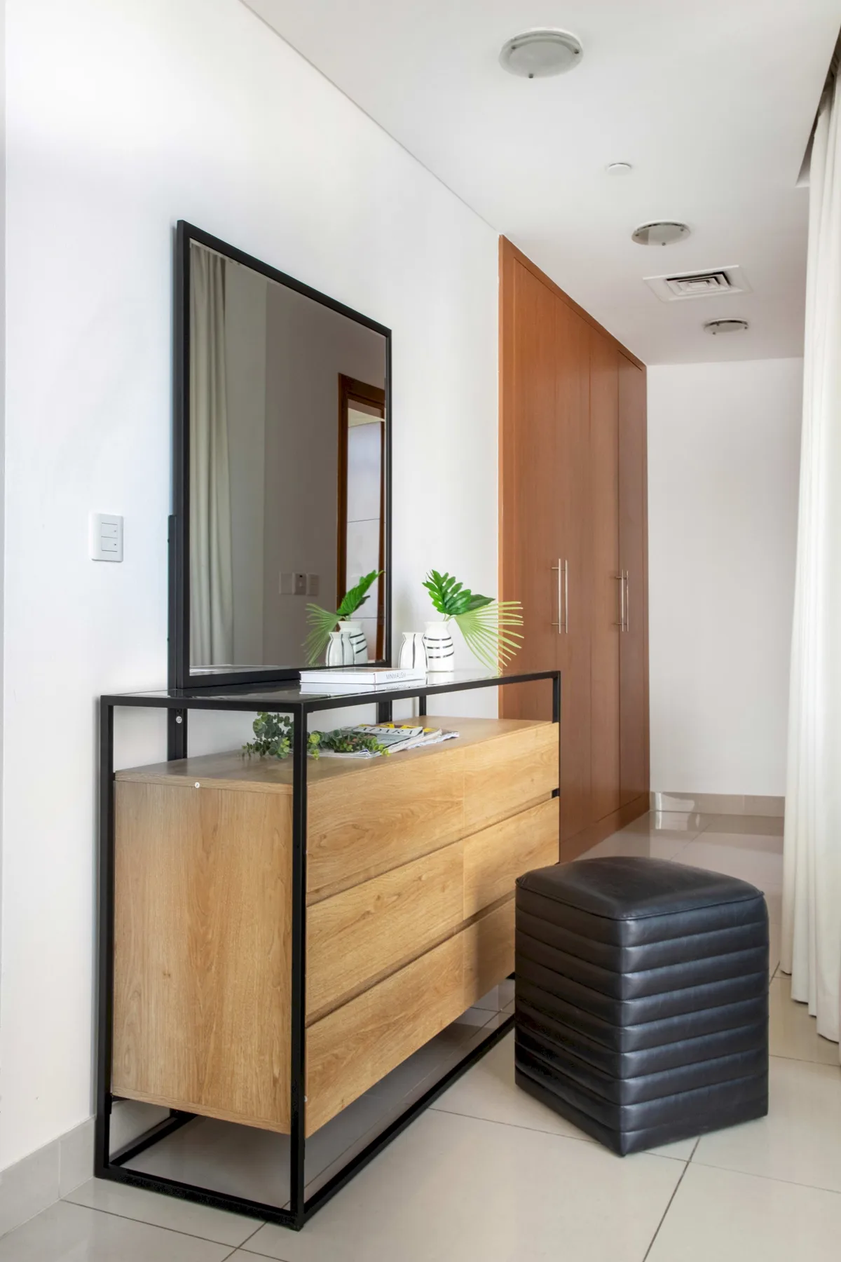 Un pasillo luminoso con una cómoda de madera, un espejo de pared y un otomano oscuro.