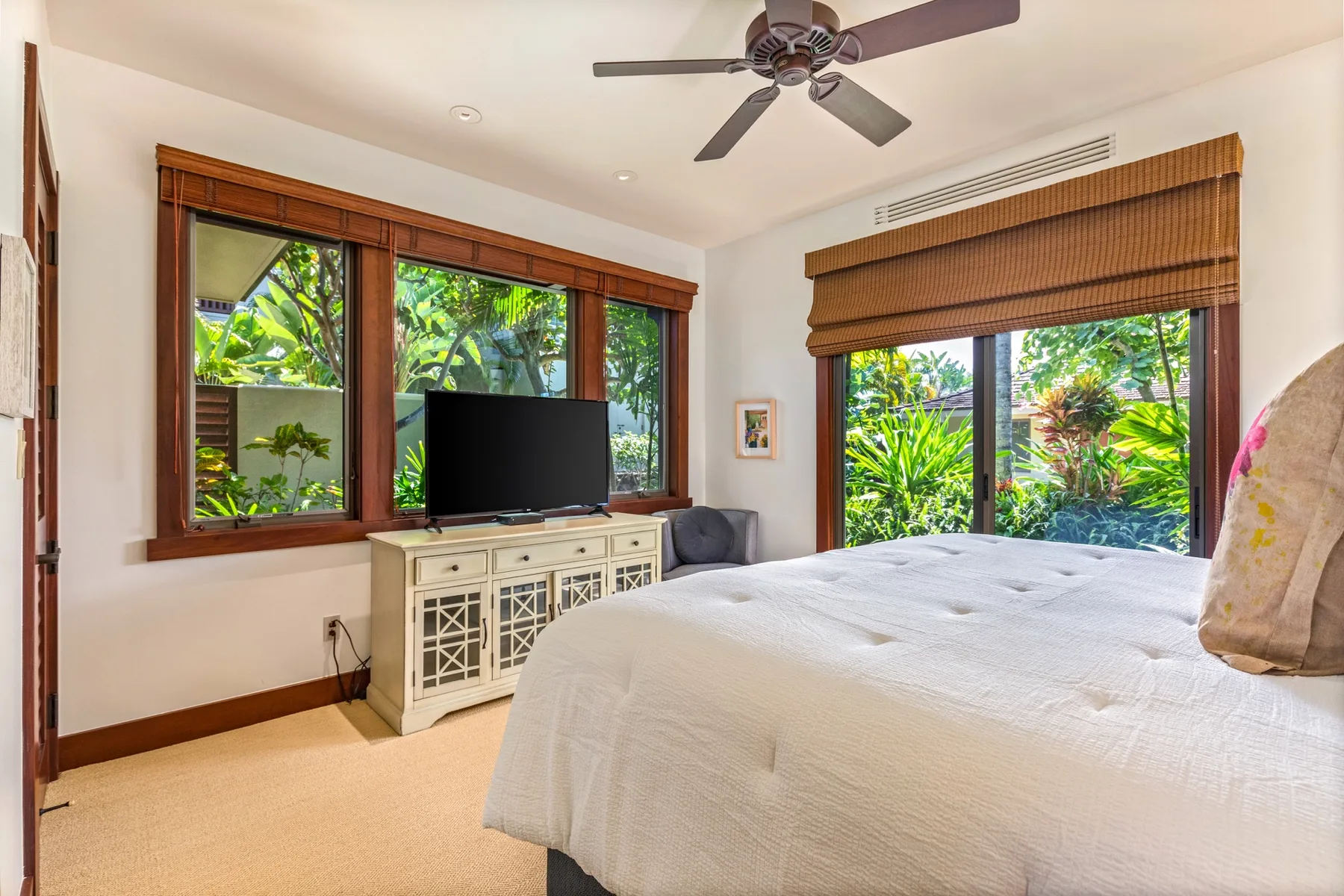 A bright bedroom with a large bed, a television, and windows overlooking greenery.