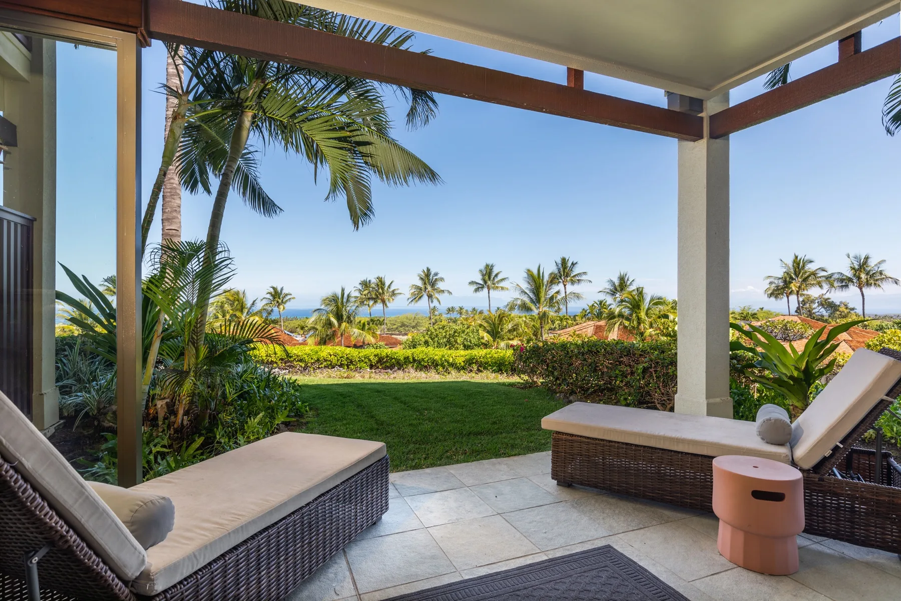 A covered patio with cushioned seating overlooks a lawn, tropical plants, and the ocean.