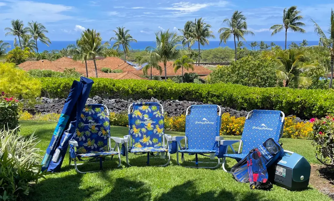 Outdoor seating and folded beach chairs sit on a lawn beside tropical landscaping.