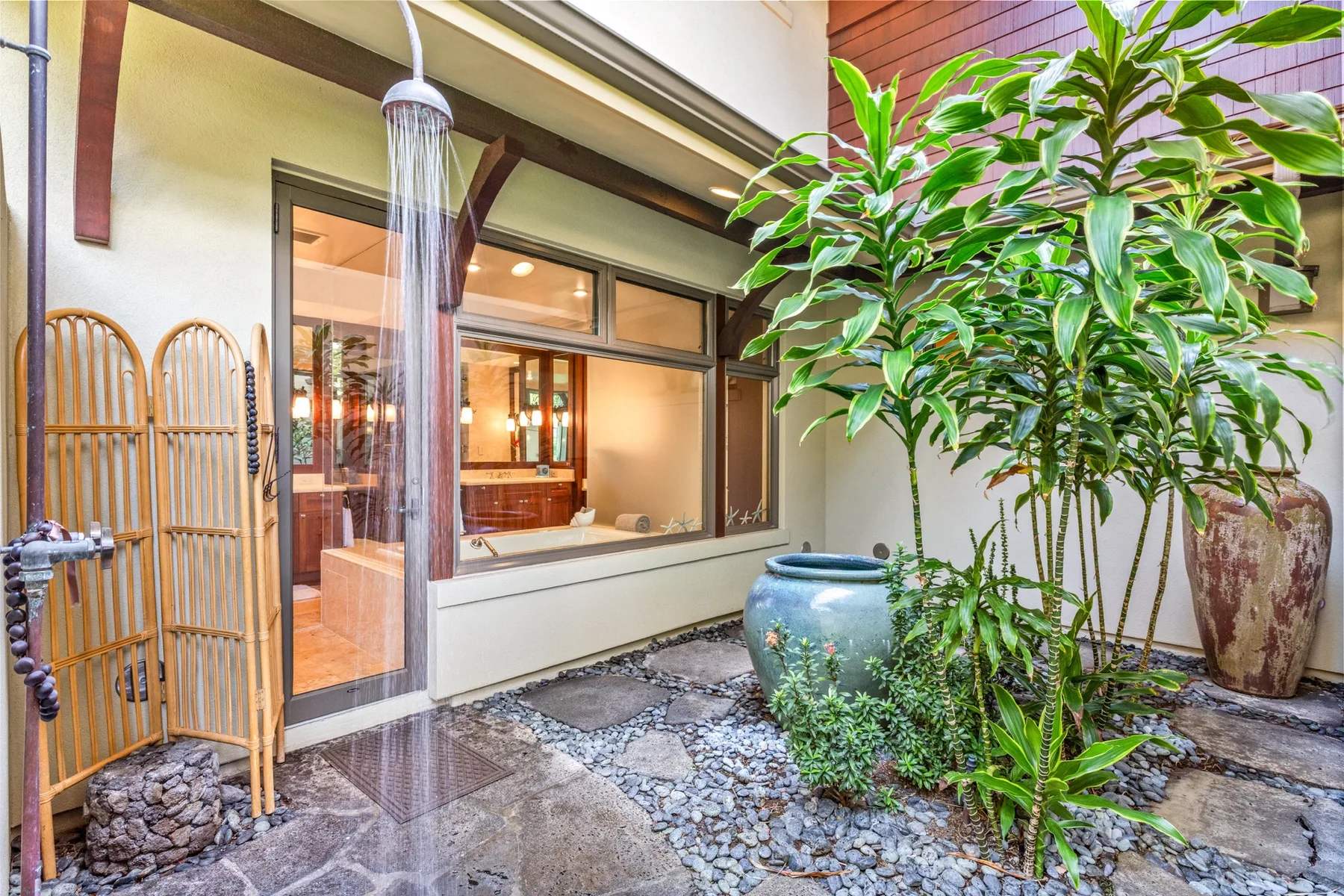 An outdoor shower area beside a large leafy plant and a window.