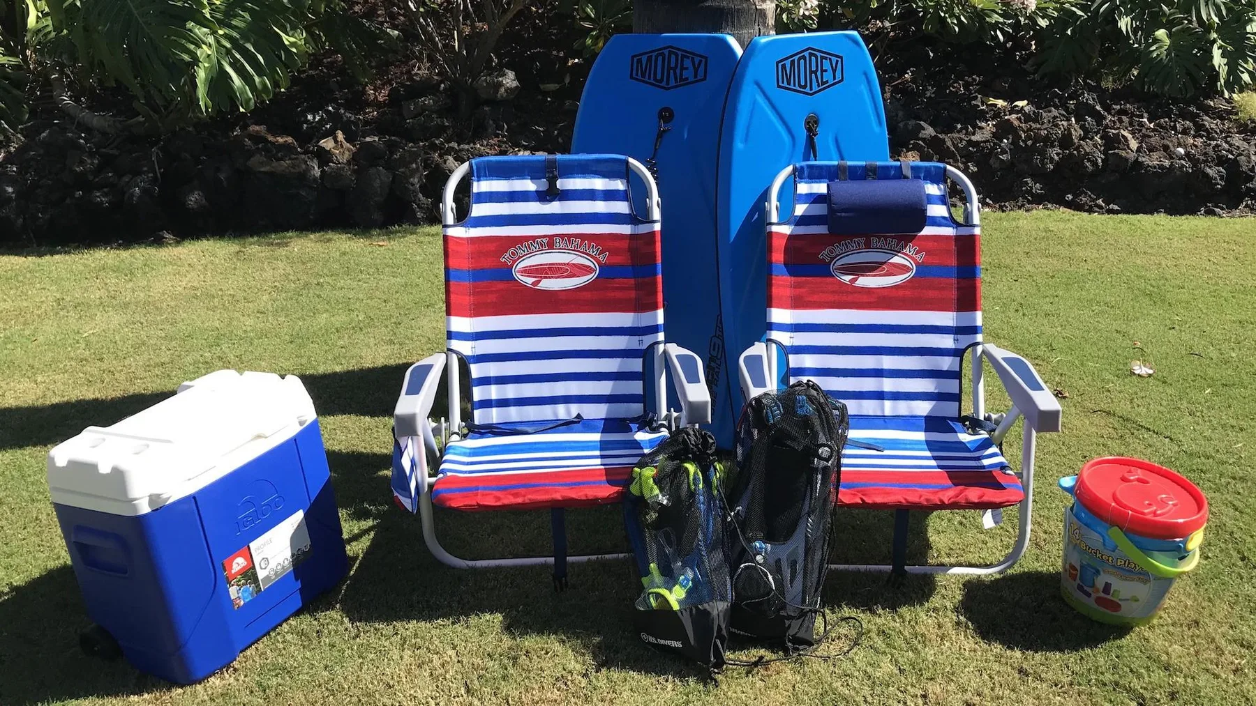 Outdoor beach gear is arranged on a grassy lawn, with striped chairs and blue bodyboards.