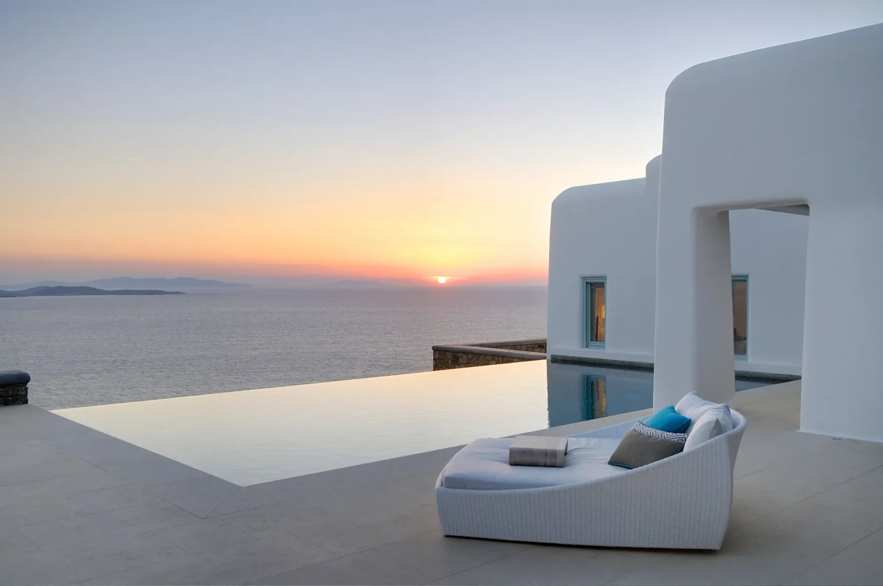 A white outdoor seating area overlooks an infinity pool and the sea at sunset.