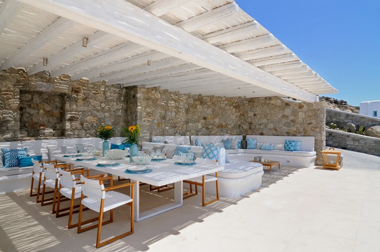 A covered outdoor dining area with tables and cushioned seating beside a stone wall.