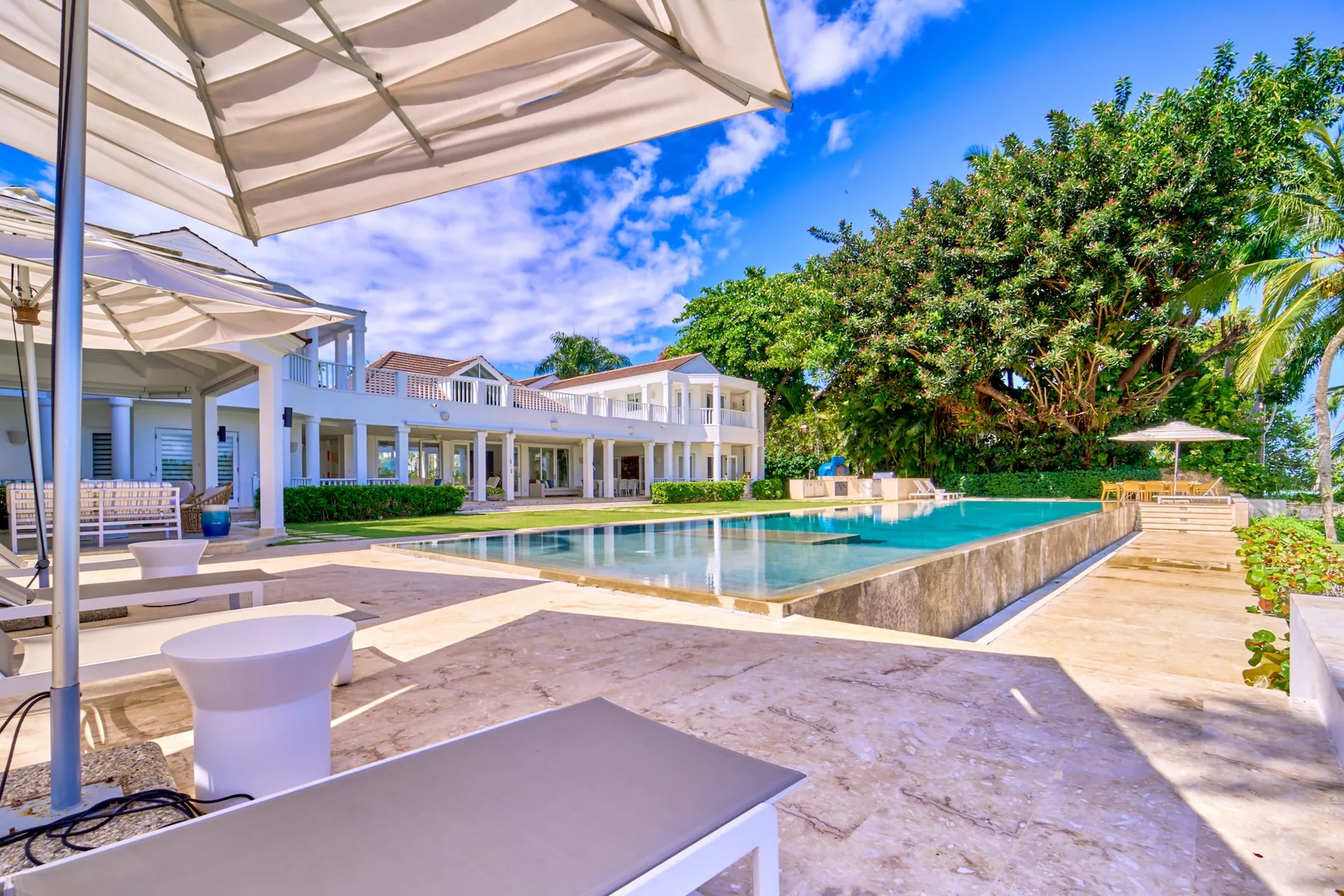 Piscina al aire libre junto a un edificio blanco, con patio sombreado y árboles frondosos.