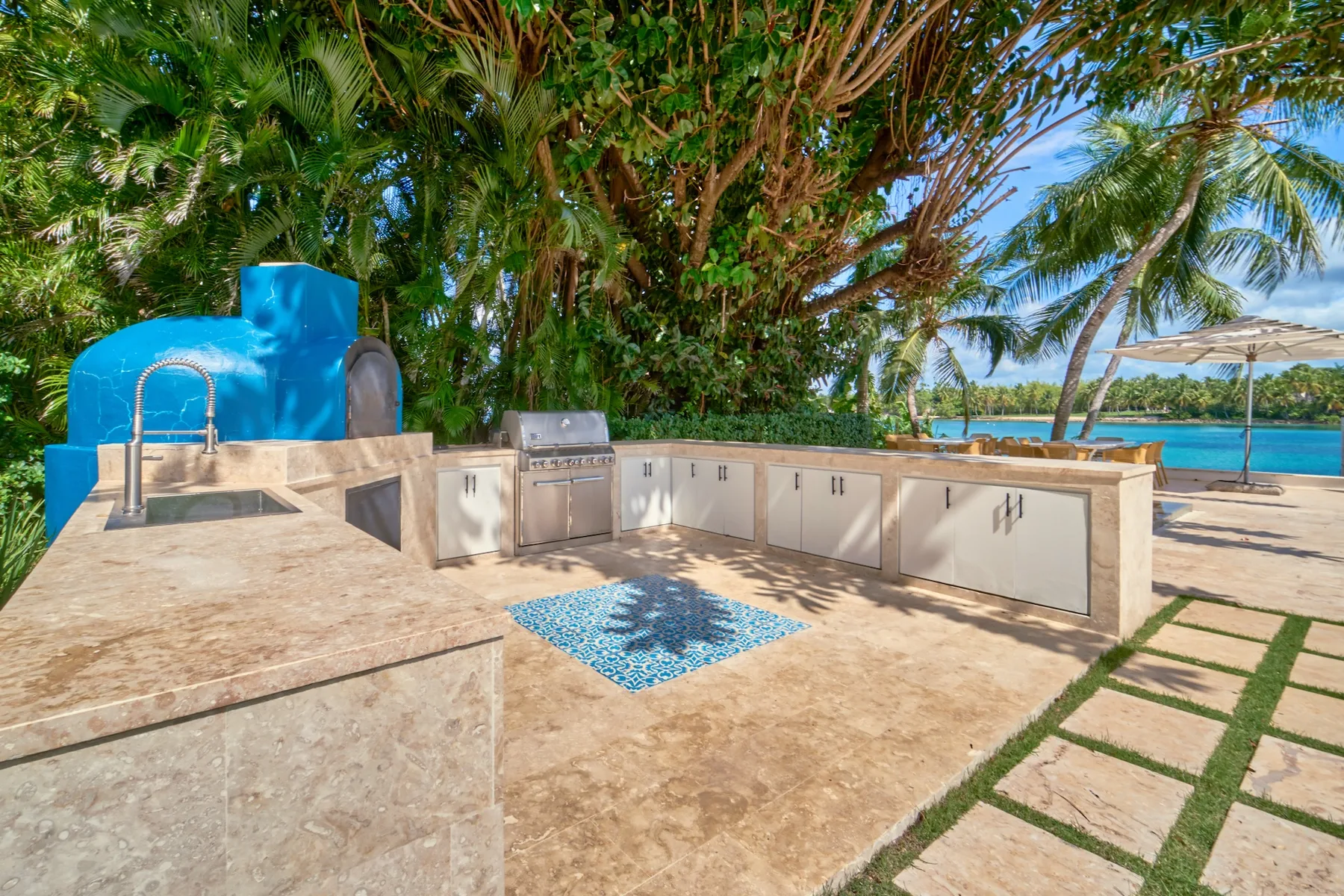 Una cocina al aire libre con encimeras y parrilla está bajo árboles tropicales junto al mar.