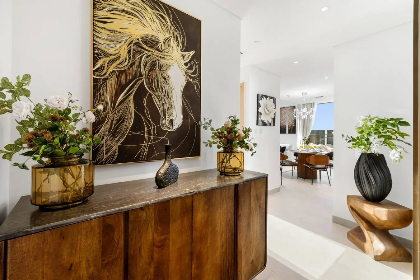 Un interior moderno y luminoso con una consola de madera, arreglos florales y una gran obra de un caballo.