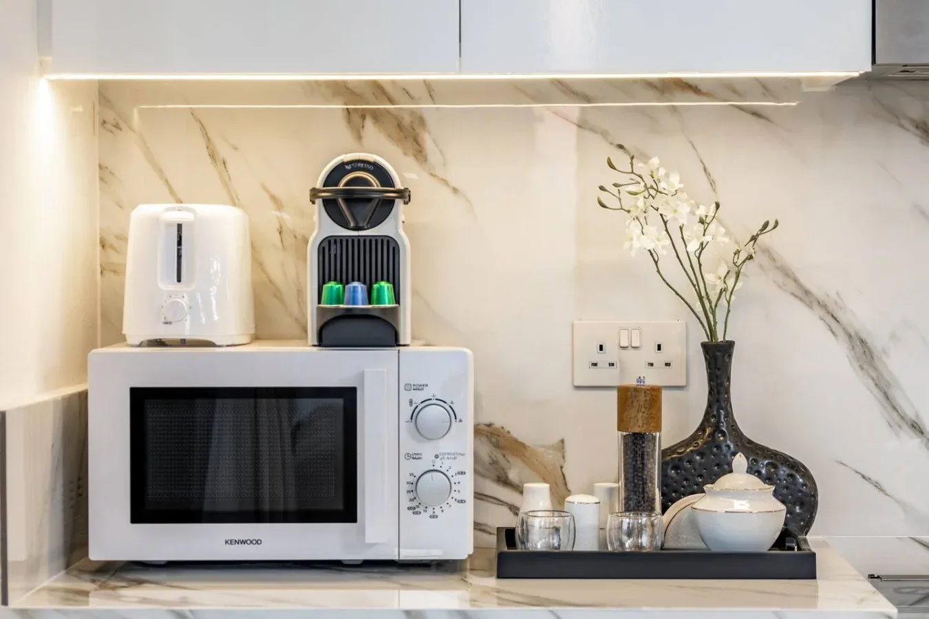 Una encimera de cocina con microondas, hervidor y cafetera bajo armarios blancos.