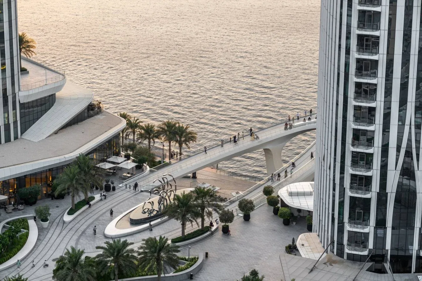 Una plaza junto al agua con senderos ajardinados y un puente peatonal.