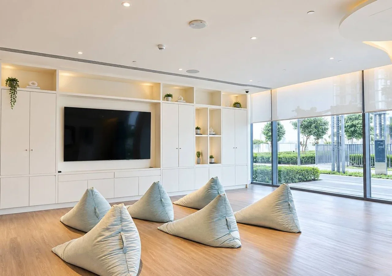 Una sala luminosa con asientos en el suelo y un televisor grande junto a amplios ventanales.