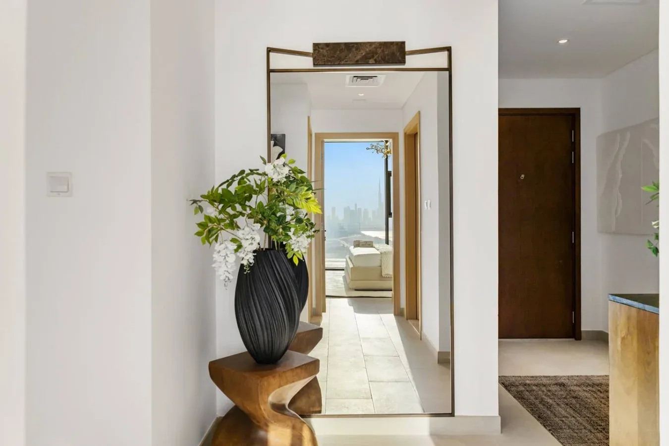 Un pasillo luminoso con un espejo, un arreglo floral y una vista hacia un dormitorio.