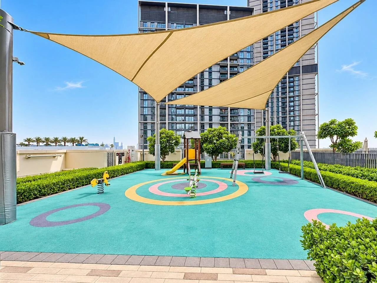 Un parque infantil colorido al aire libre está bajo grandes toldos, con un edificio alto detrás.