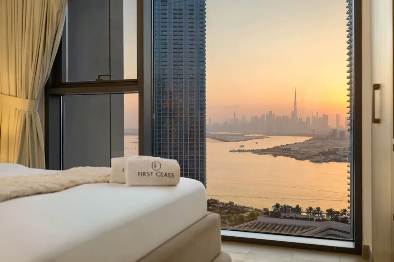 Un dormitorio con una cama tendida frente a un río y un horizonte urbano al atardecer.