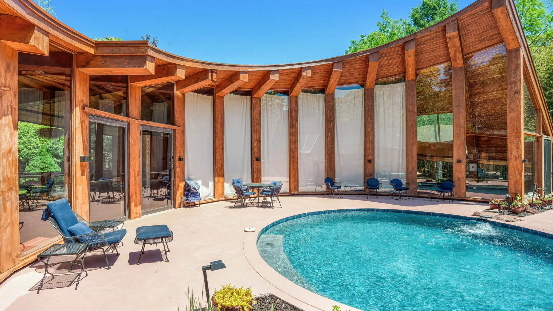 Una piscina exterior curva está junto a un edificio con estructura de madera y asientos en el patio.