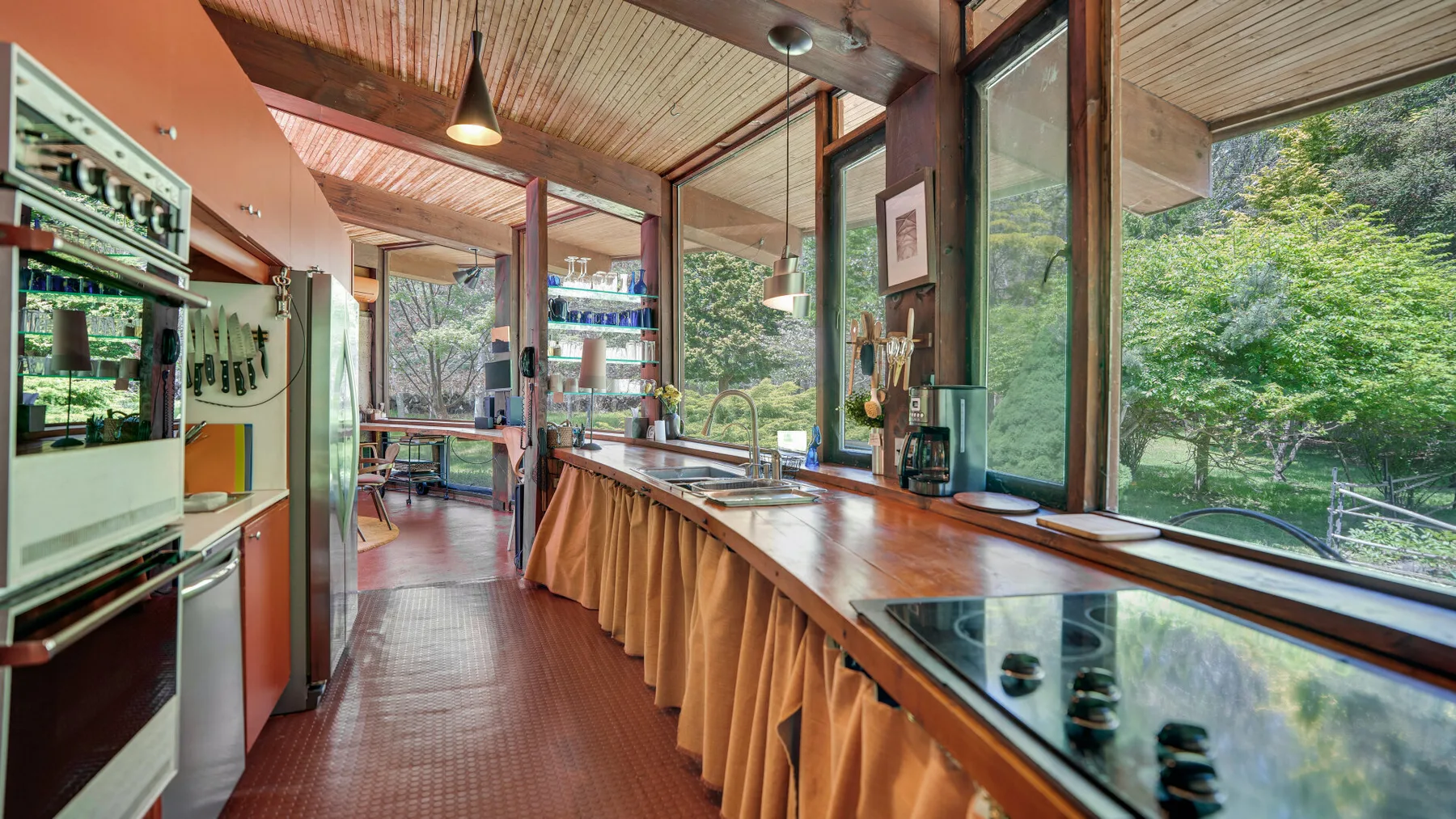 Una encimera de cocina se extiende bajo grandes ventanas con vistas a árboles verdes.