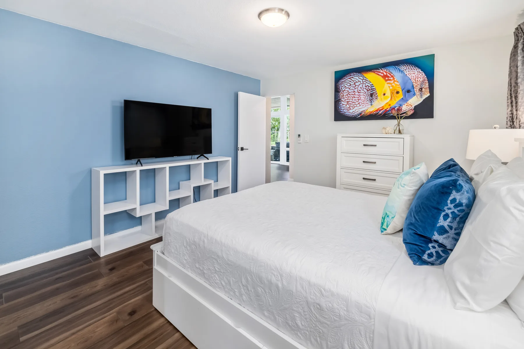 Dormitorio con cama blanca, pared azul y televisor sobre un mueble bajo.