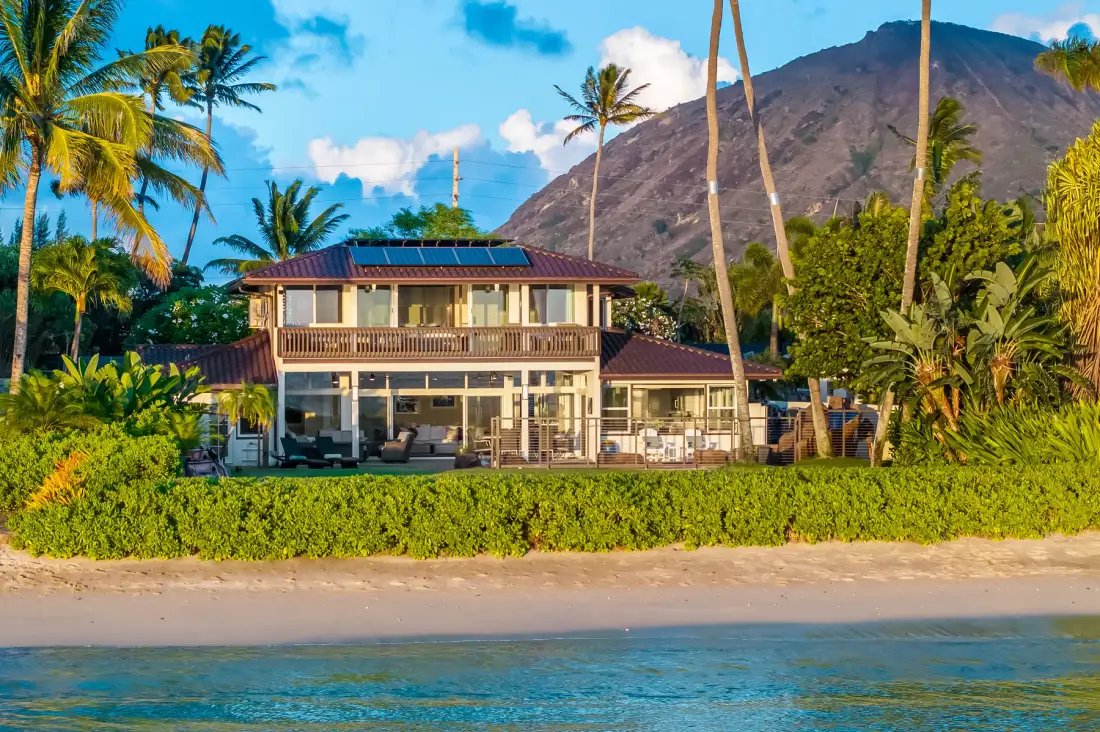 Una casa frente a la playa con una galería cubierta está rodeada de palmeras tropicales, bajo una montaña.