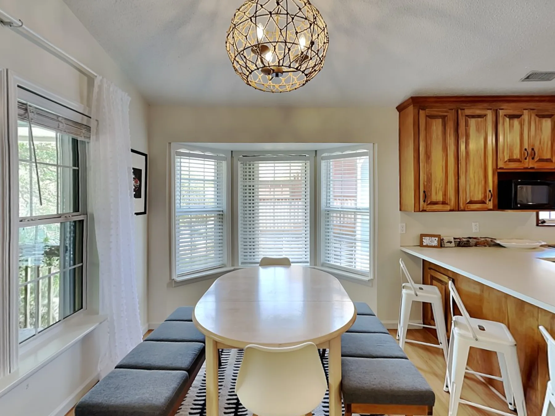 Un comedor luminoso tiene una mesa larga junto a una ventana y armarios de cocina.