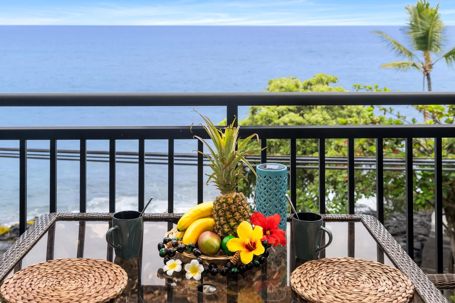 Una mesa de balcón con frutas tropicales y flores mira al océano.