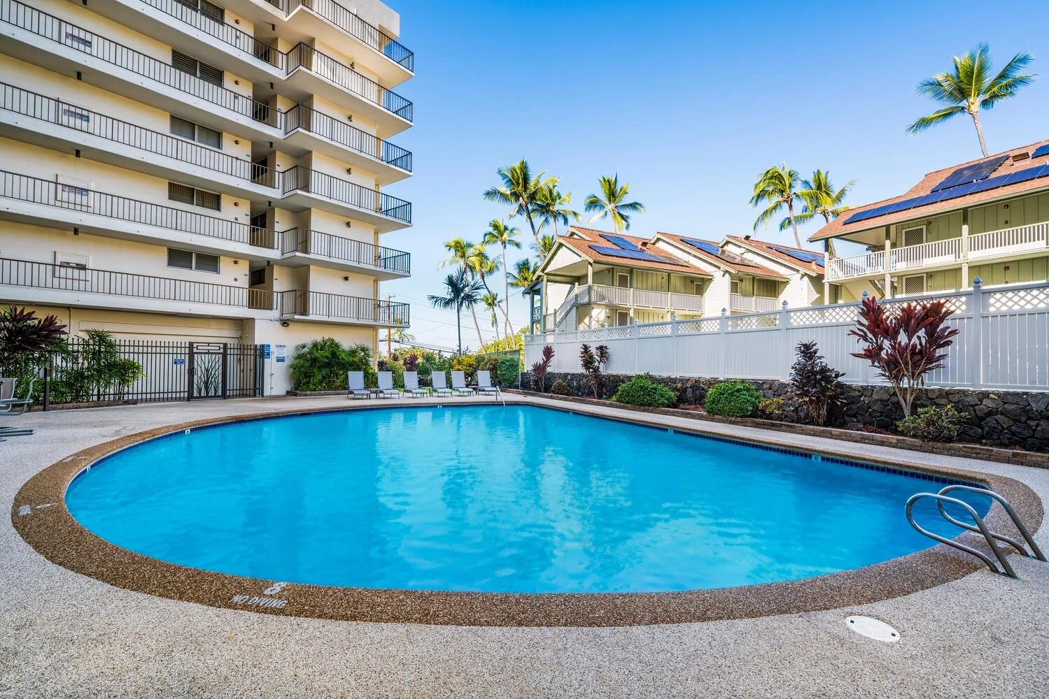 Una piscina exterior curva está junto a un edificio de varios pisos y vegetación tropical.