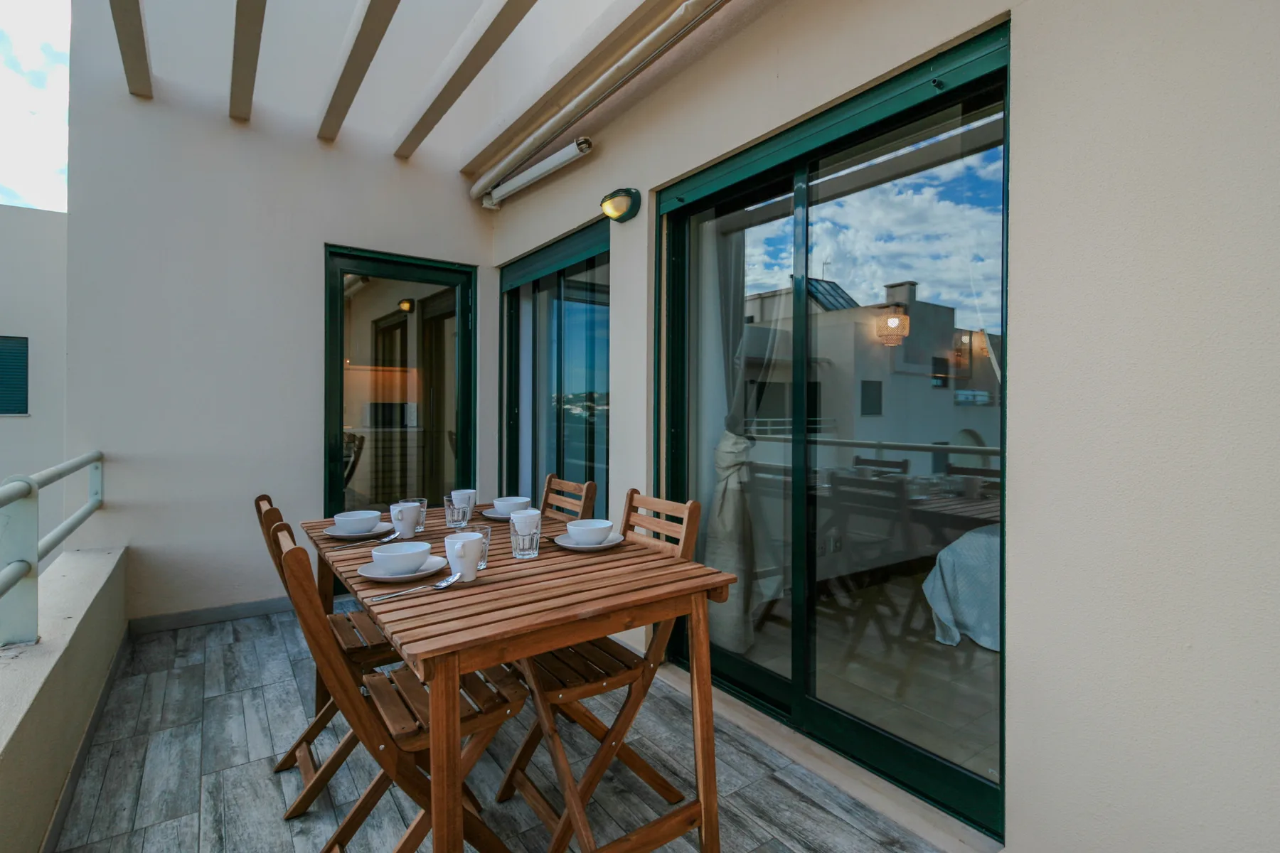 A covered balcony has a wooden dining table and seating beside sliding glass doors.