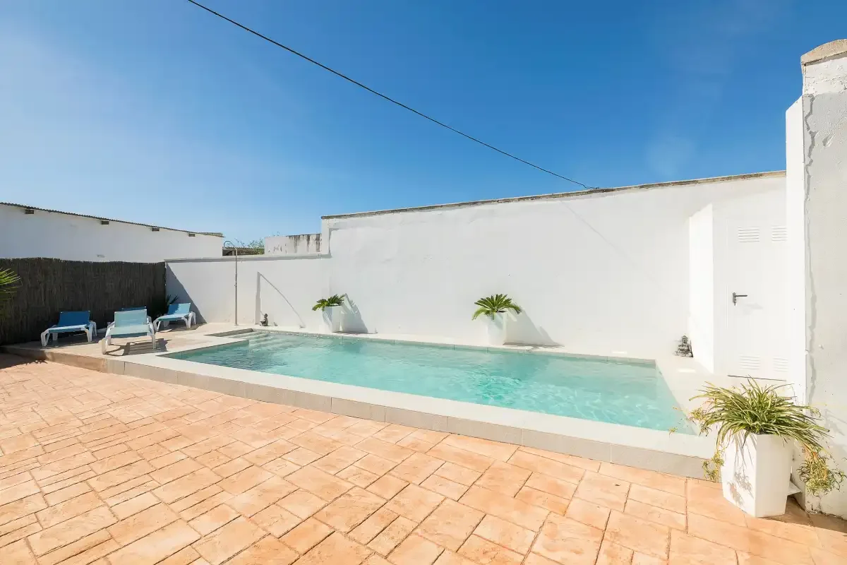 An outdoor swimming pool sits beside a white-walled patio with lounge seating.