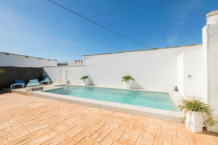 An outdoor pool sits beside a white wall and a sunlit paved patio.