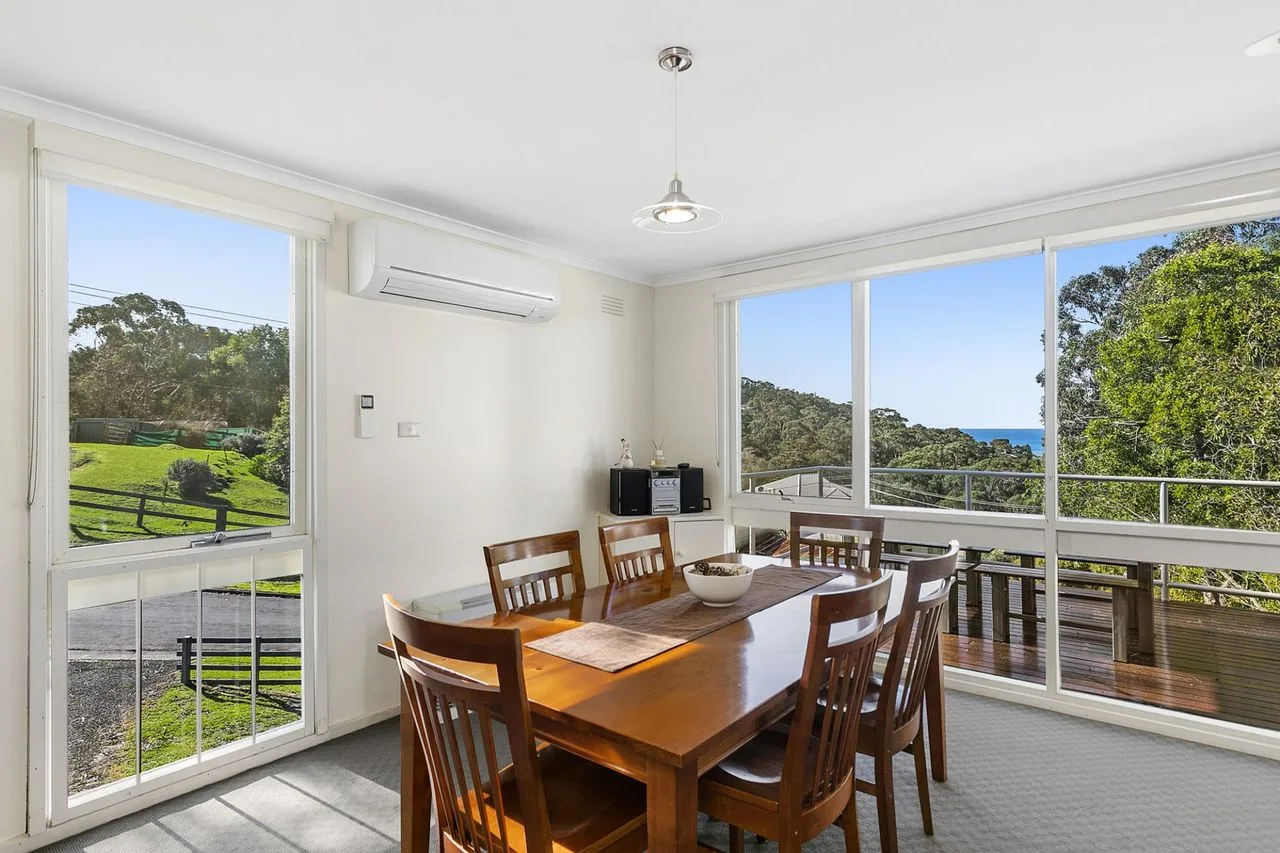 A dining area with a wooden table and large windows overlooking greenery.