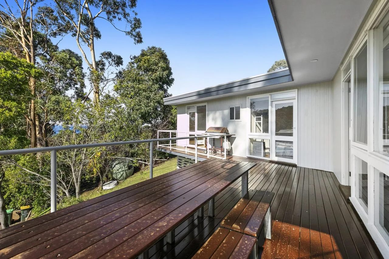 An outdoor deck with a long wooden table beside white walls and trees.