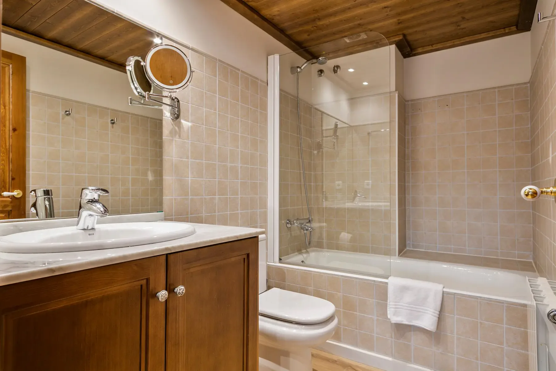 A tiled bathroom with a wooden vanity, sink, toilet, and bathtub with a glass screen.