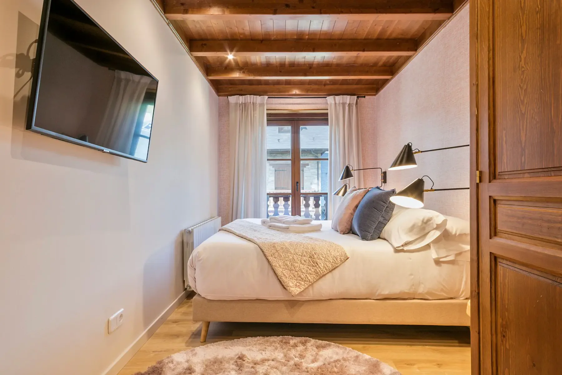 A bedroom with a bed beneath exposed wooden ceiling beams and a curtained window.