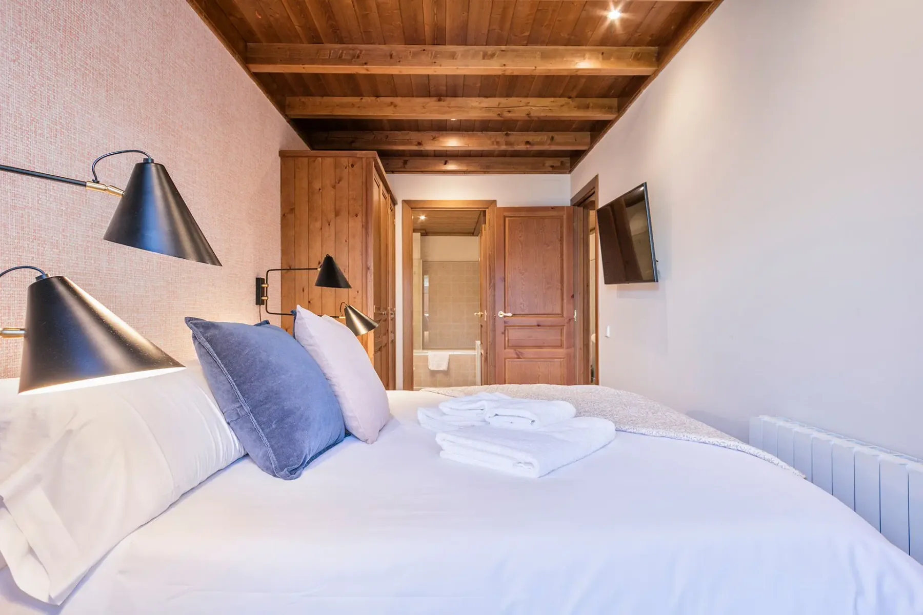 A bedroom with white bedding, blue pillows, and exposed wooden ceiling beams.