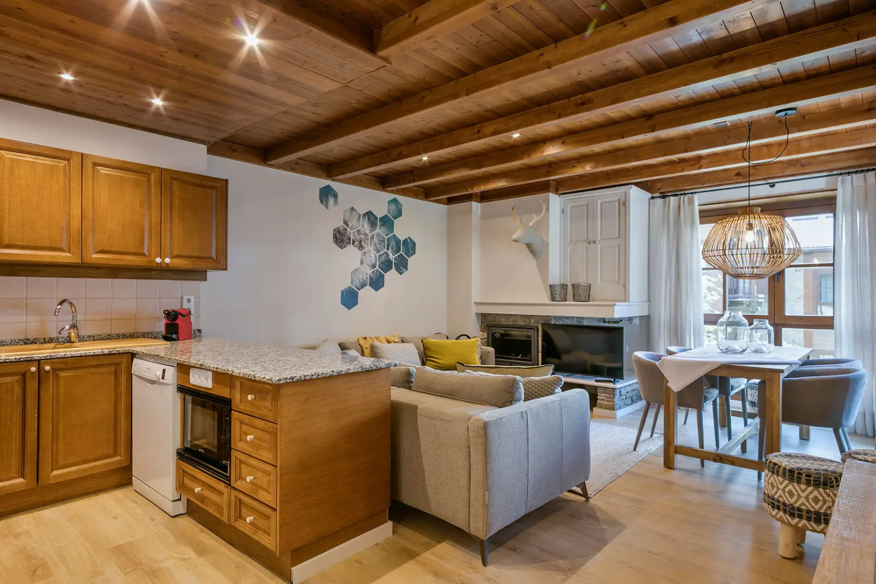 A modern open-concept living and dining area with wooden accents. A gray sofa, wooden coffee table, and poufs are in the foreground, while a dining table with chairs and kitchen are in the background. Large windows provide natural light.
