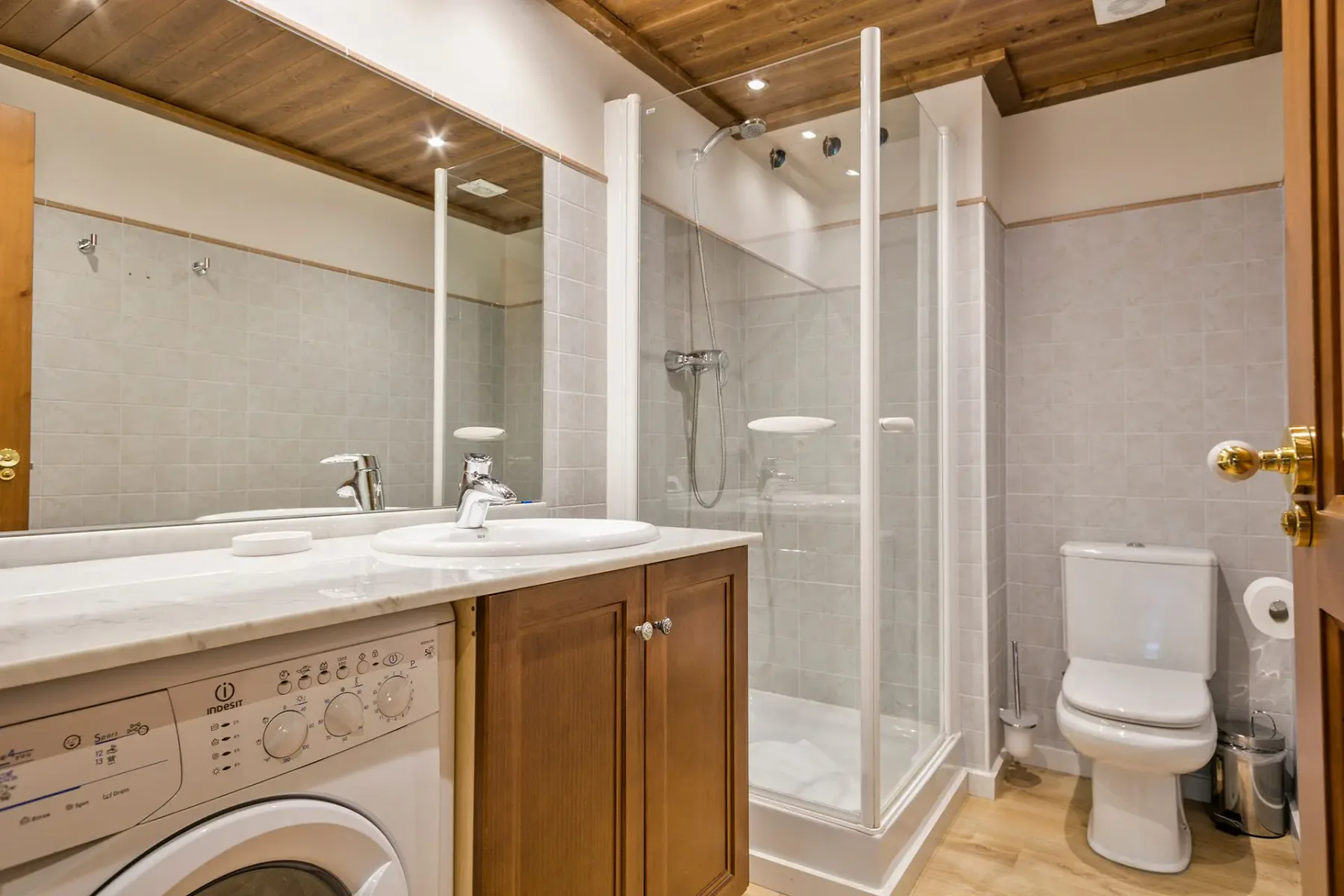 Modern bathroom with wooden accents, a vessel sink on a wooden vanity, a wall-mounted toilet, a bathtub with a glass enclosure, and a large window.