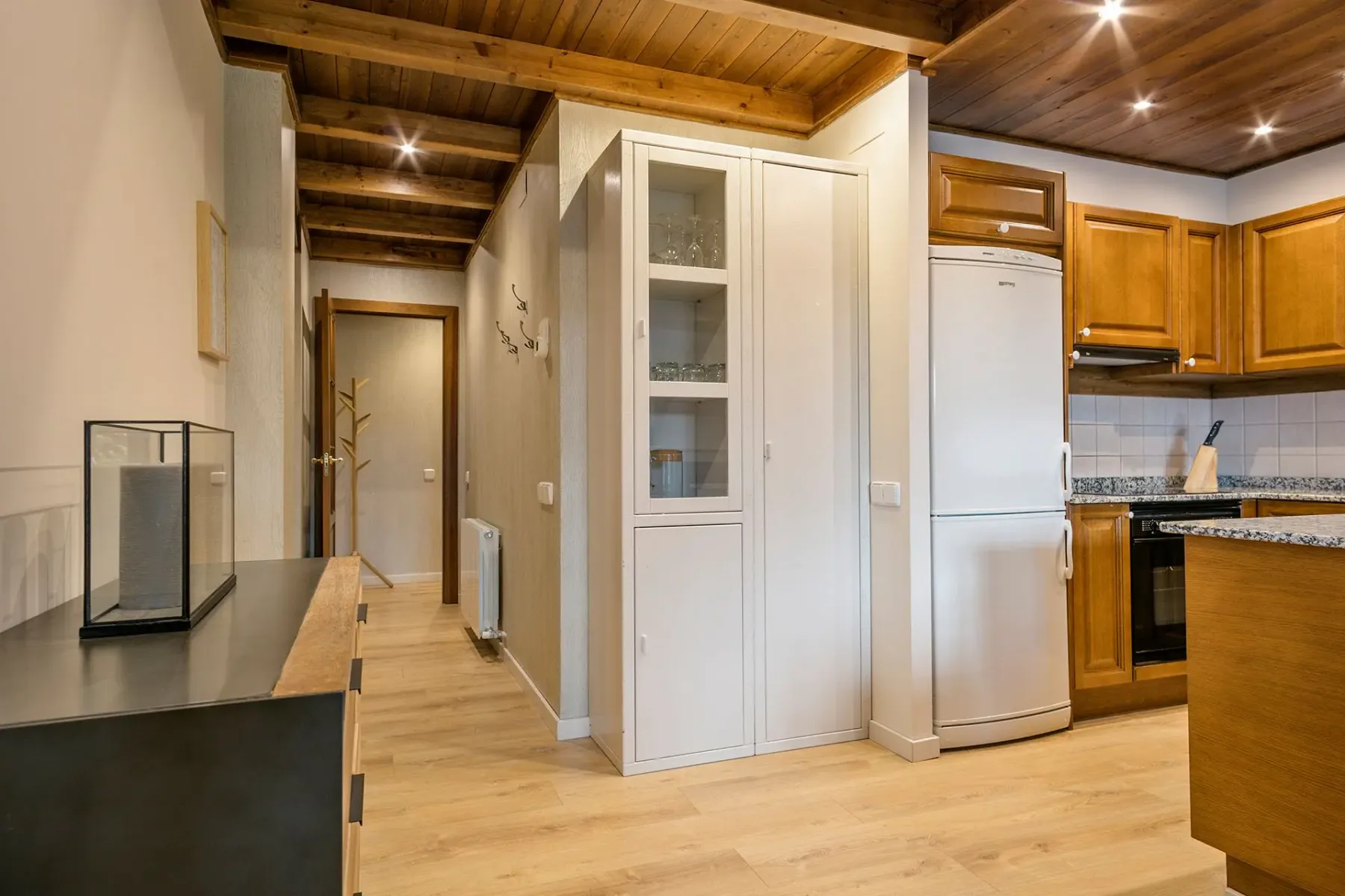 A kitchen with wooden cabinets, a refrigerator, and a hallway beyond.