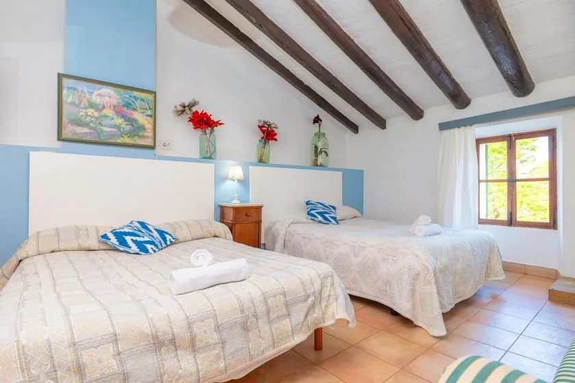 A bright bedroom with two beds, exposed ceiling beams, and a sunlit window.