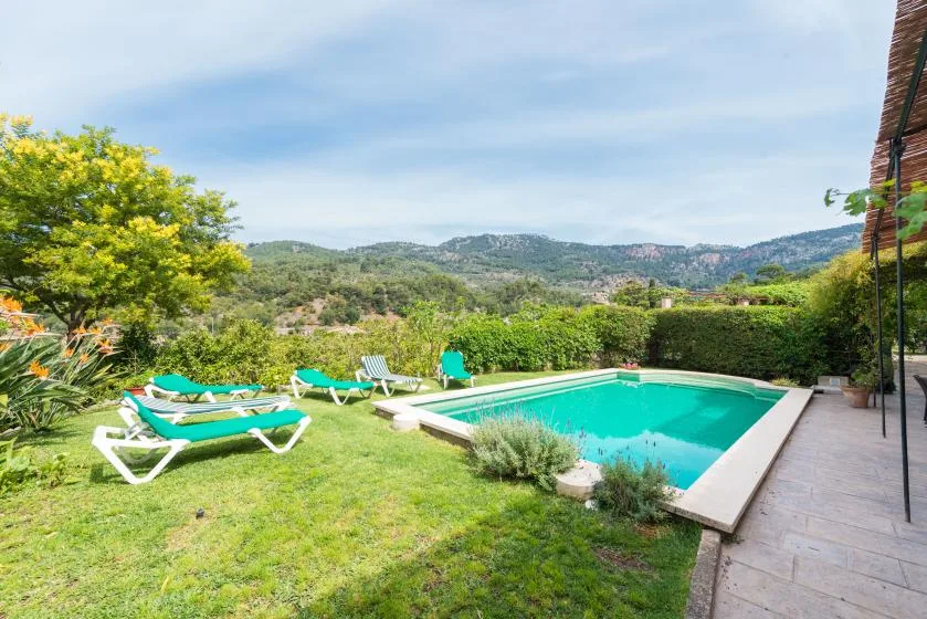 An outdoor swimming pool sits beside a grassy yard with loungers and hills beyond.