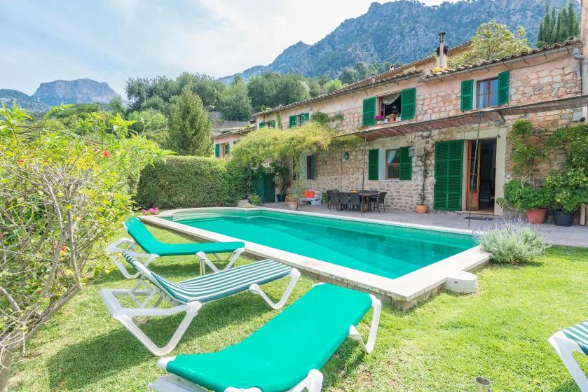 A swimming pool and sun loungers sit beside a stone house in a mountain landscape.