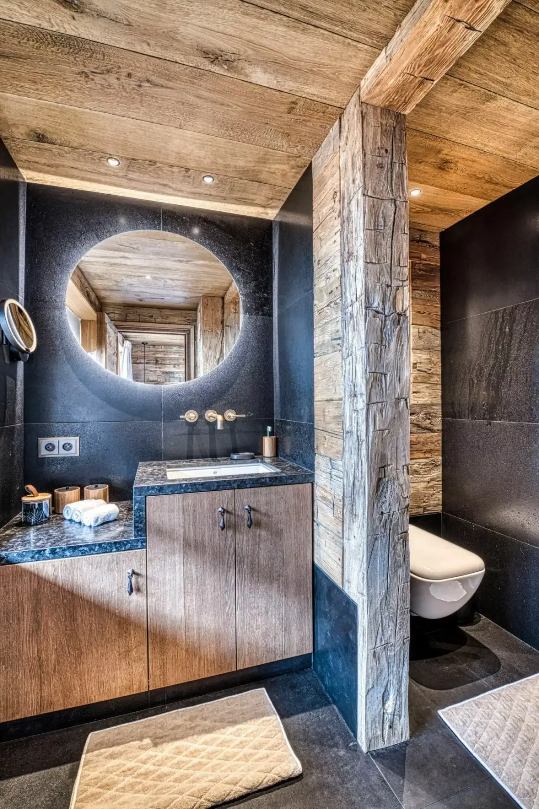 A bathroom vanity with a large round mirror and a toilet behind a partition.